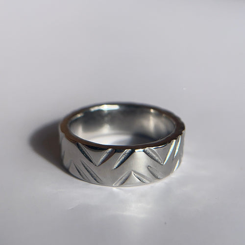 A silver band ring with a textured chevron or zigzag pattern, resting on a light gray surface with soft shadows.
