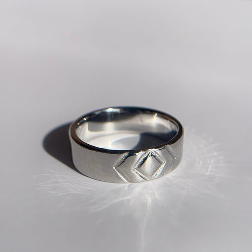 A silver-toned geometric band ring with a diamond-shaped cutout detail, resting on a white surface under direct sunlight.