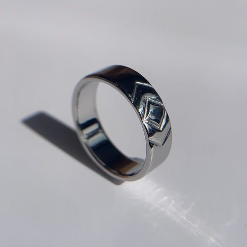 A silver band ring with a geometric, faceted chevron-like pattern on the outer surface, photographed on a white surface with a hard shadow.
