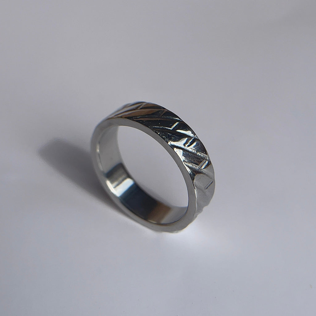 A silver metallic band ring with a textured, geometric patterned exterior, resting on a plain light gray surface.