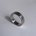 A silver metallic band ring with a textured, geometric patterned exterior, resting on a plain light gray surface.