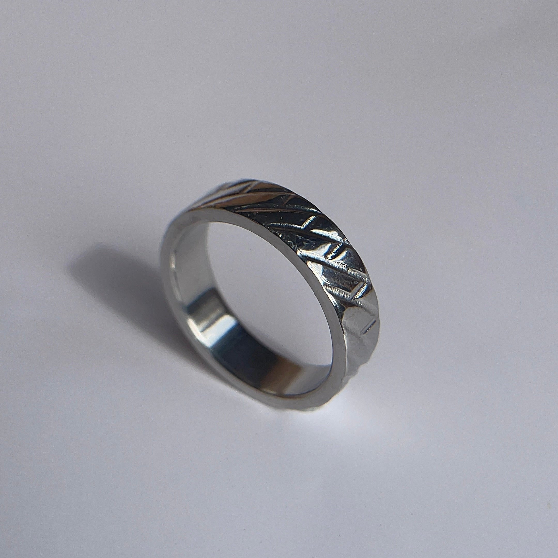 A silver metallic band ring with a textured, geometric patterned exterior, resting on a plain light gray surface.
