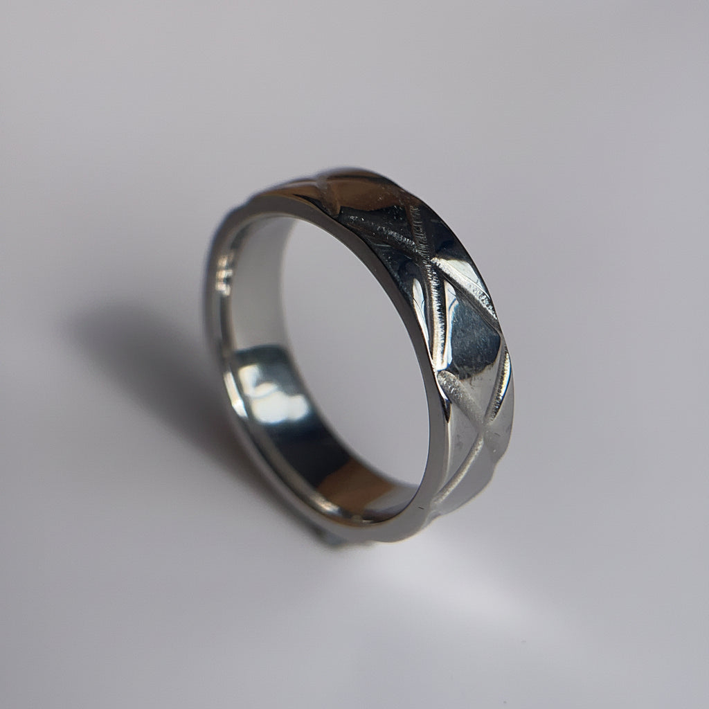 A silver-toned metal ring with a textured, crisscrossed pattern on its outer band, resting on a plain light gray surface.