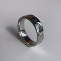 A silver-toned metal ring with a textured, crisscrossed pattern on its outer band, resting on a plain light gray surface.