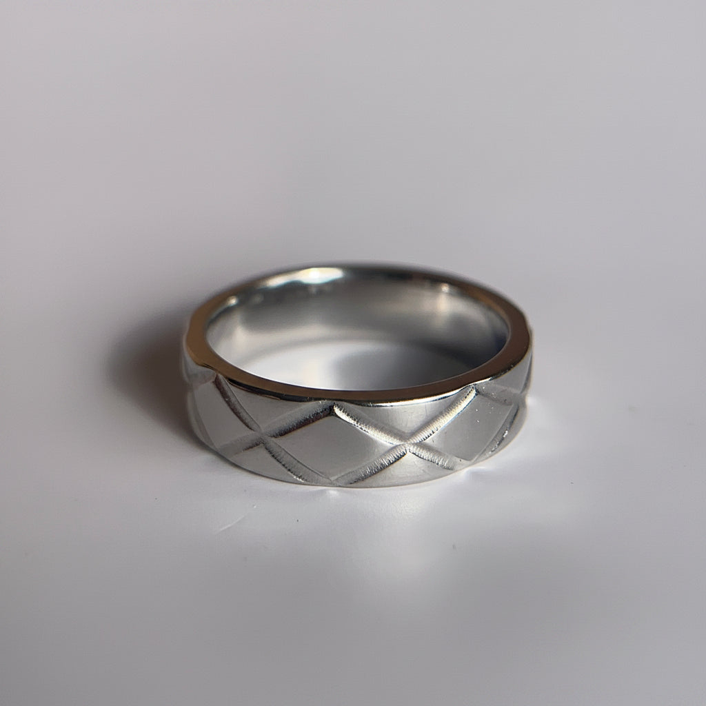 A silver band ring with a textured, diamond-grid pattern on the exterior, resting on a plain light gray surface.