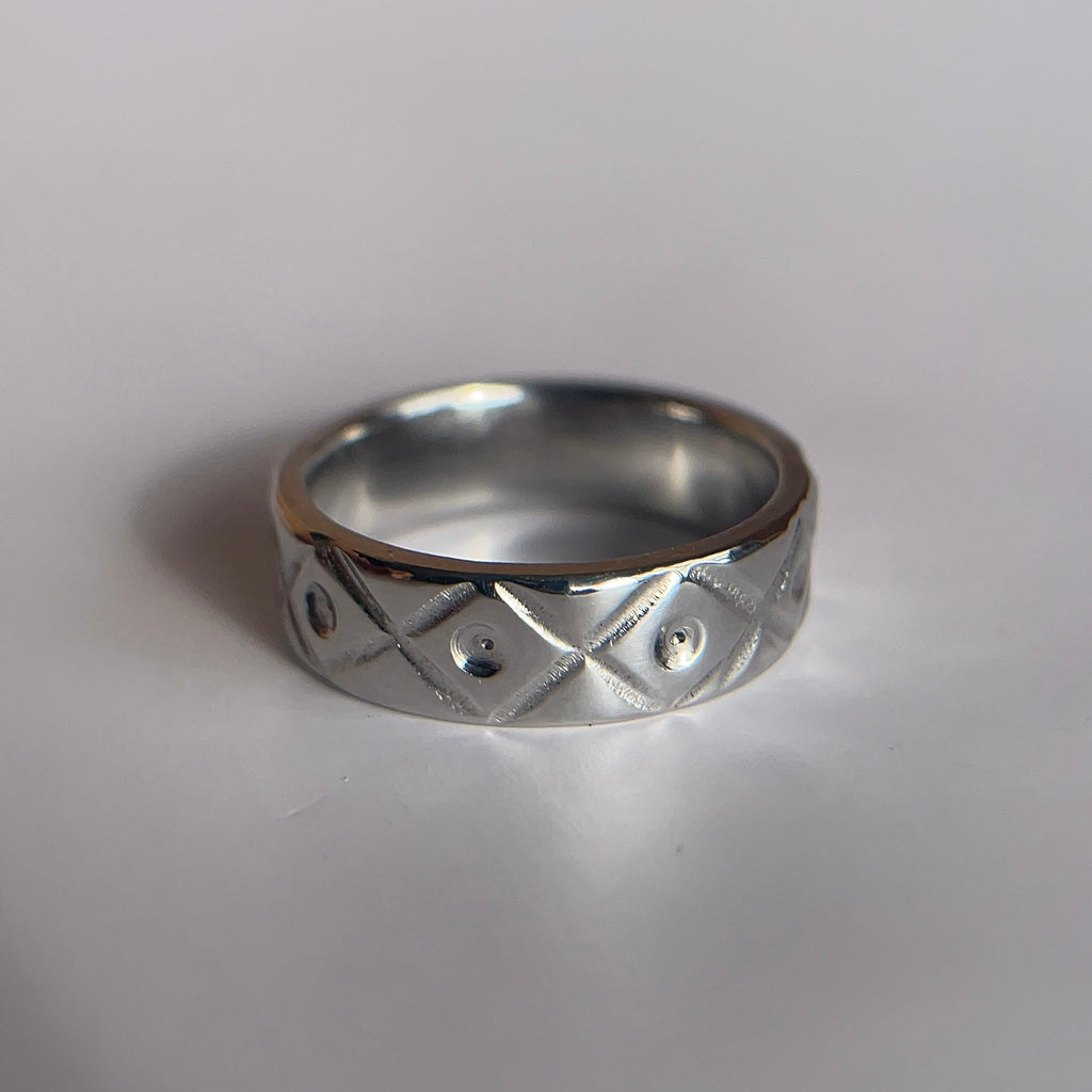 A silver band ring with a textured geometric diamond pattern, resting on a plain light gray surface.