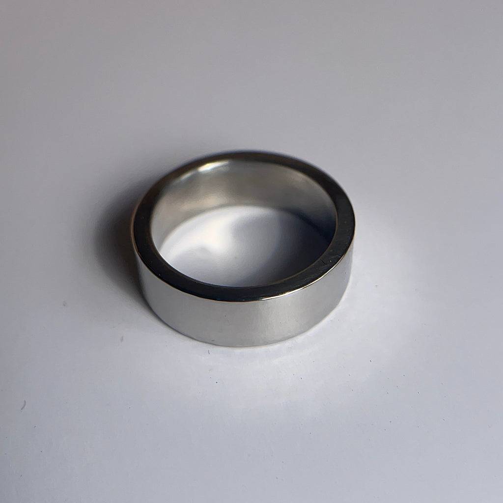 A simple silver-toned metal band ring, likely titanium, resting on a plain white surface with a soft shadow.