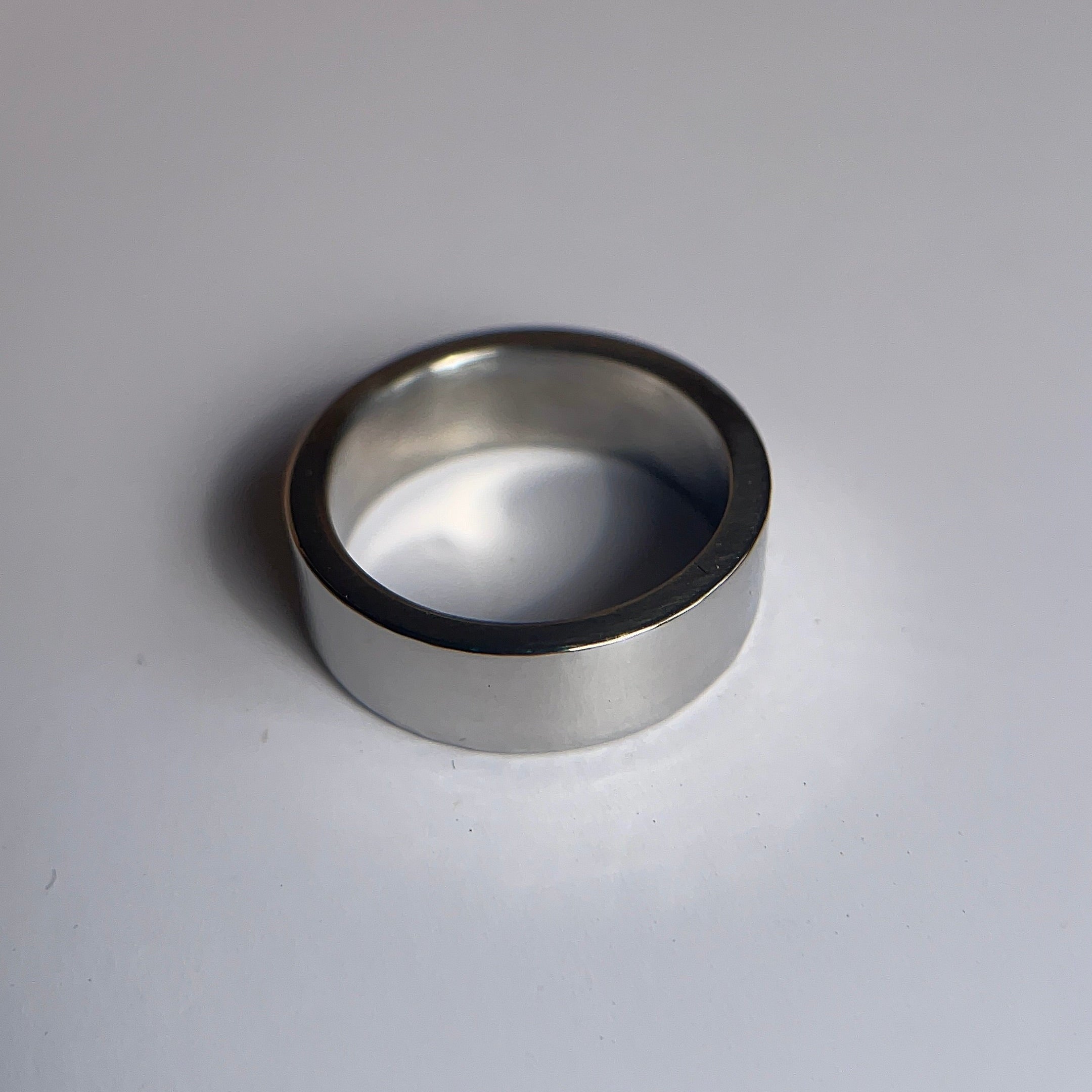 A simple silver-toned metal band ring, likely titanium, resting on a plain white surface with a soft shadow.