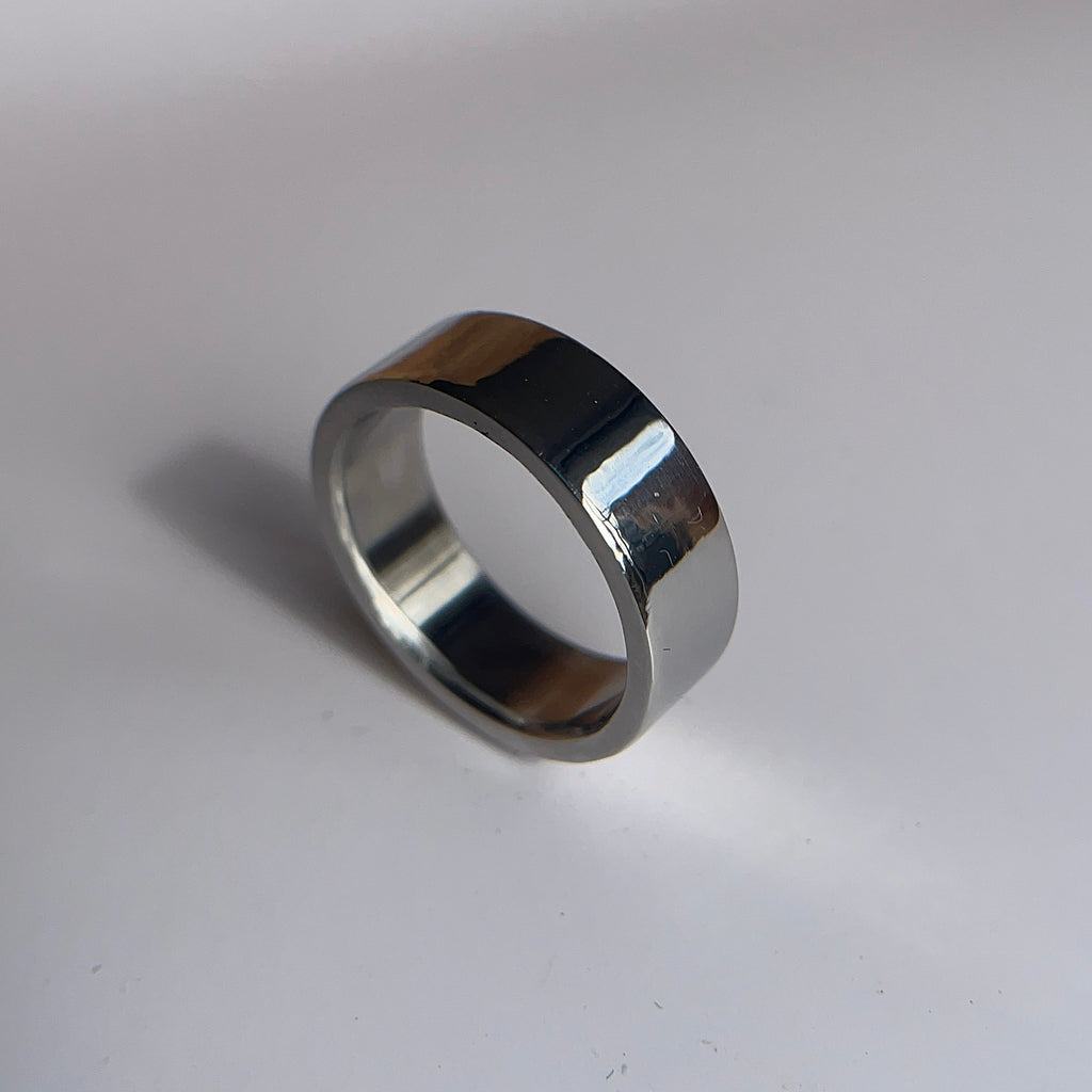 A polished silver metal band ring standing upright on a light gray surface with a soft shadow.
