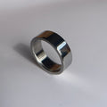 A polished silver metal band ring standing upright on a light gray surface with a soft shadow.