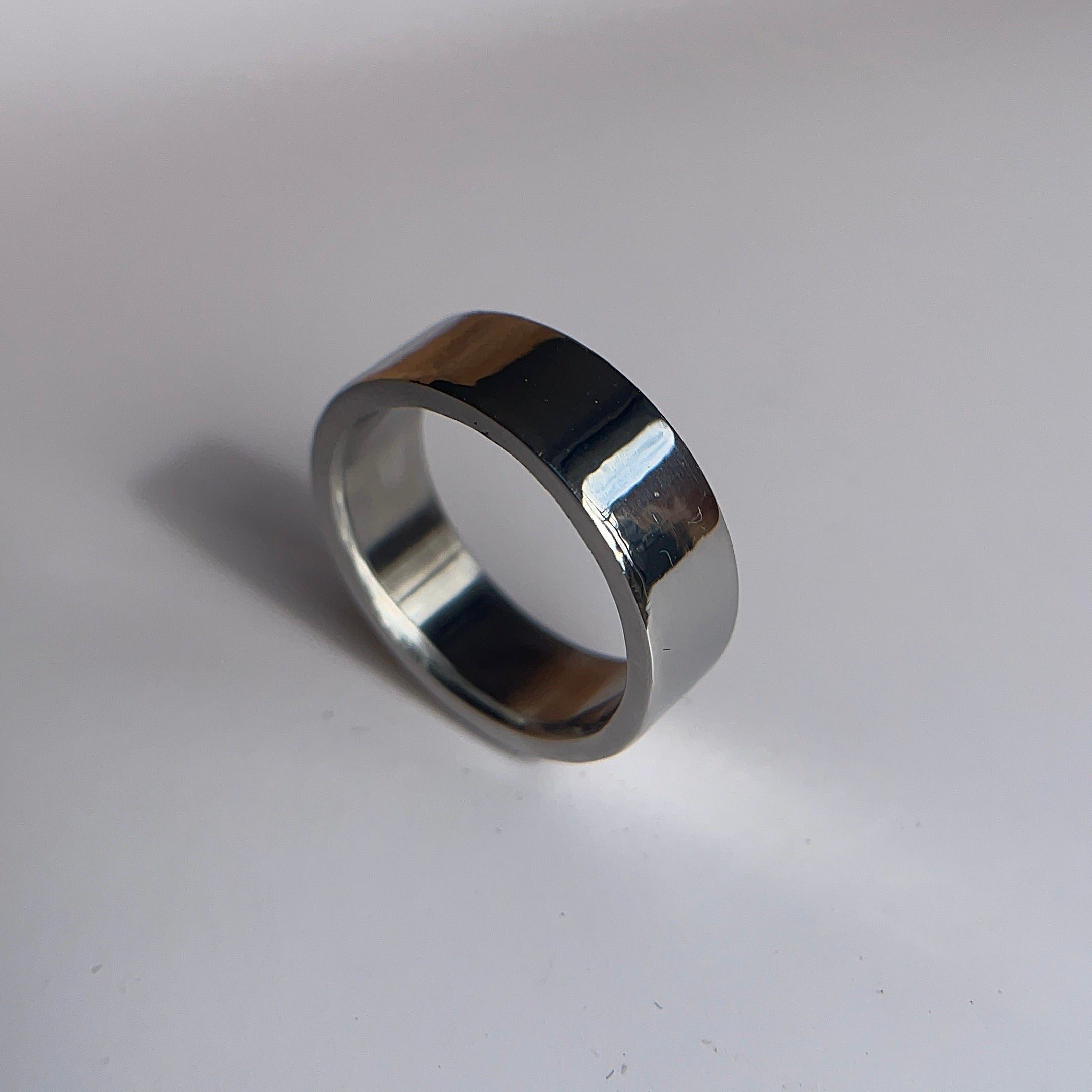 A polished silver metal band ring standing upright on a light gray surface with a soft shadow.