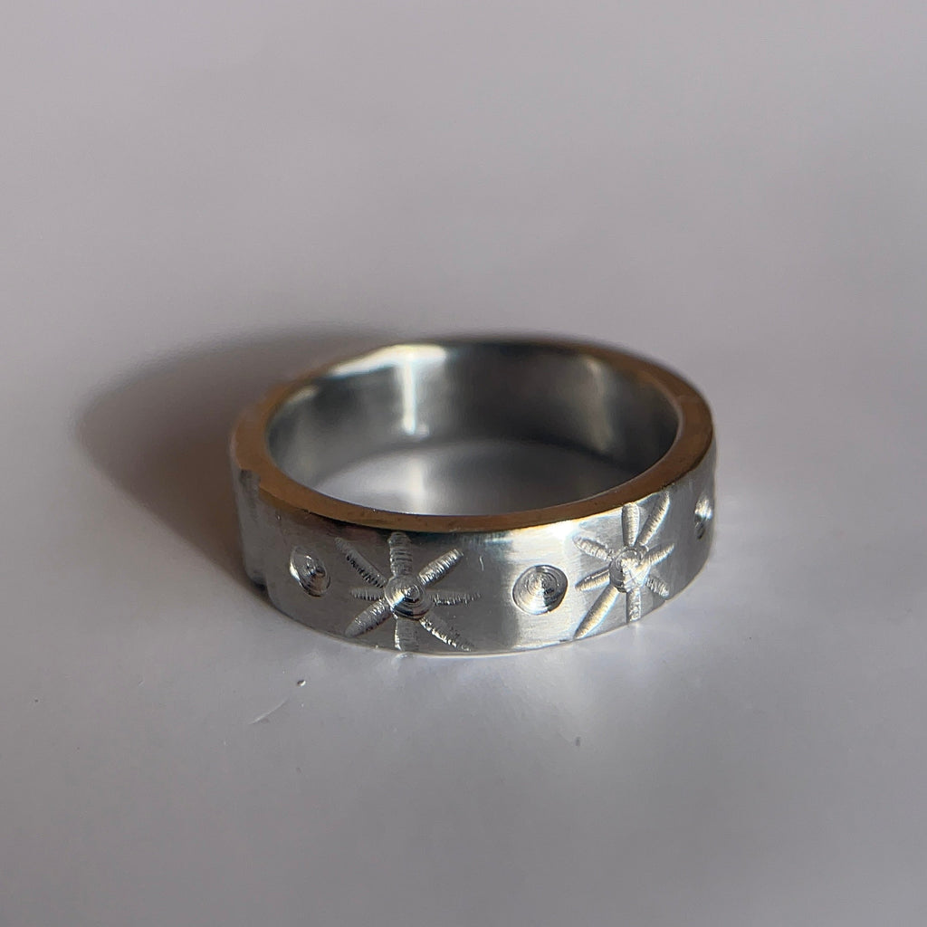 A silver-toned metal ring with engraved starburst and circular cutout patterns, resting on a plain light gray surface.